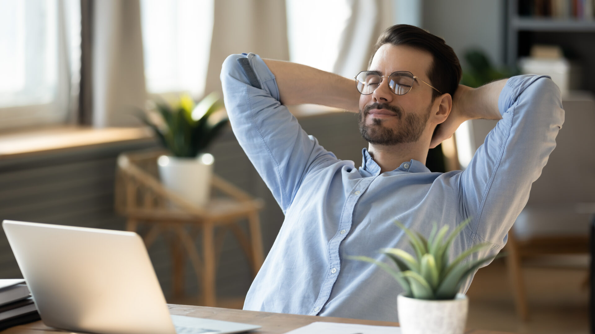 Calm millennial man in glasses sit relax at home office workplace take nap or daydream. Happy relaxed Caucasian young male rest in chair distracted from computer work, relieve negative emotions.